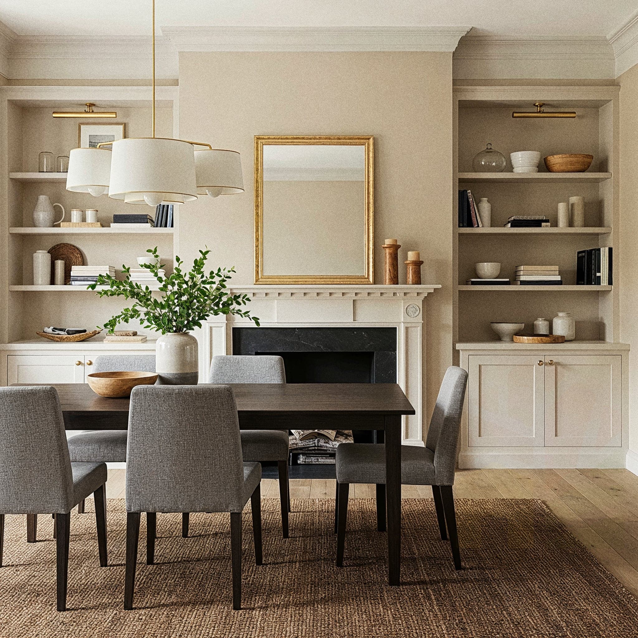 A nostalgic dining room with a Farmhouse Rustic design, featuring a weathered wooden dining table with a checkered table runner, surrounded by wooden chairs with cushioned seats in muted red. The room includes a vintage chandelier with warm bulbs, a distressed wooden sideboard with a mason jar centerpiece and dried flowers, and a braided jute rug. Large windows with sheer curtains allow natural light to highlight the dark wooden floor and whitewashed walls, creating a warm and inviting dining atmosphere.
